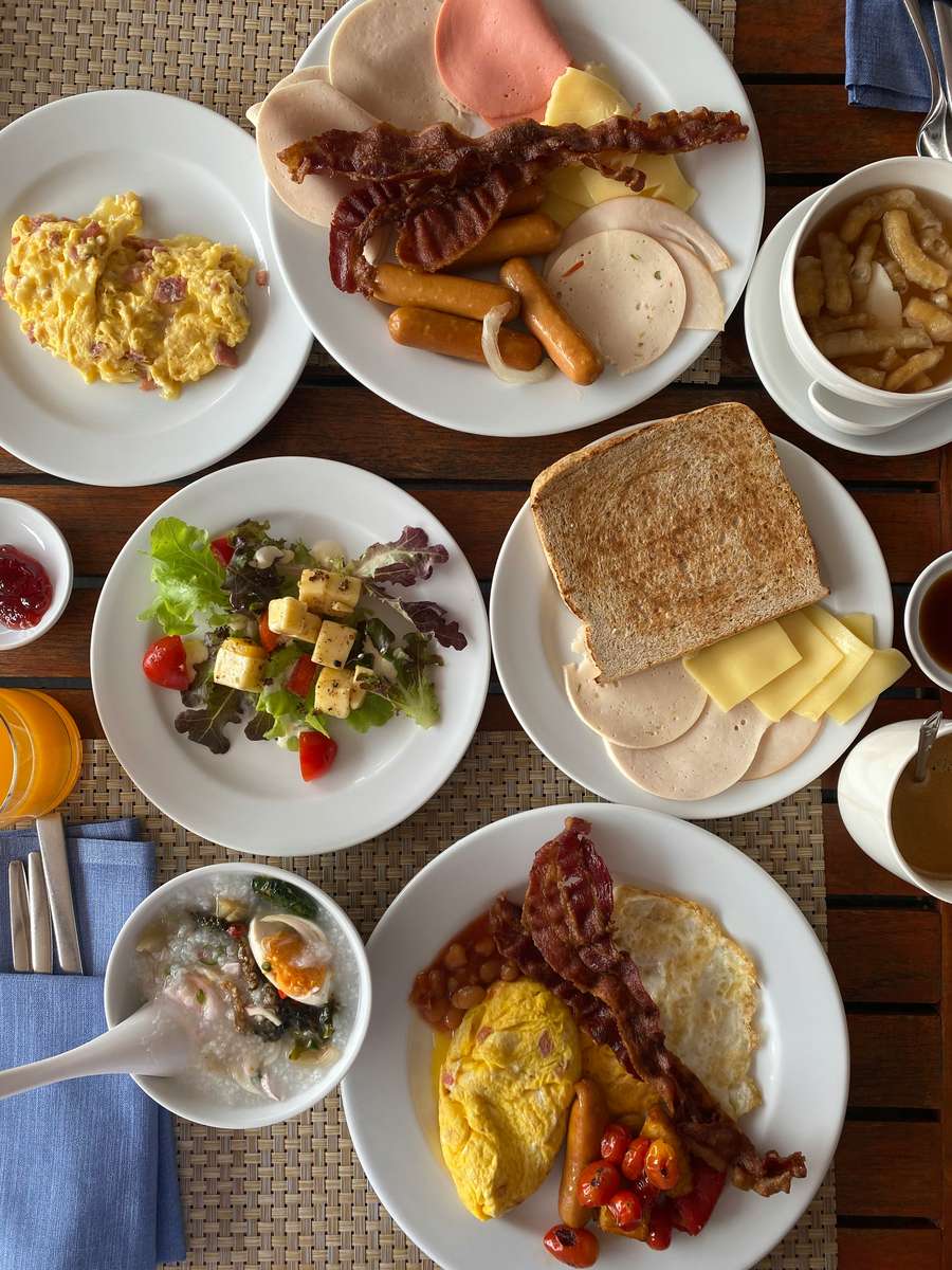 Overhead view of a large breakfast spread with eggs, bacon, sausages, sliced meats and cheese, toast, salad, rice porridge, soup, and drinks.