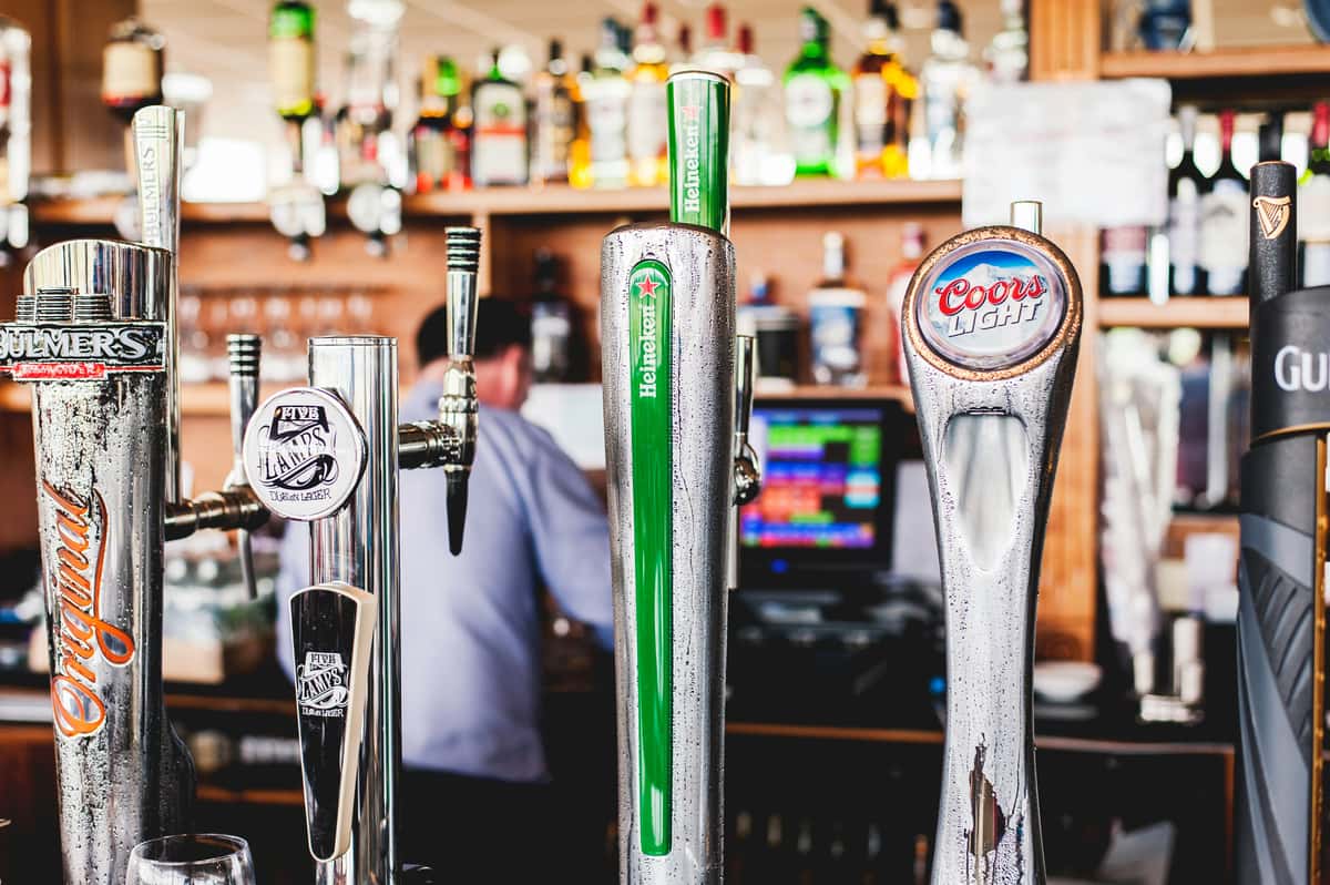 Assorted beer taps featuring Heineken, Coors Light, Five Lamps, and Bulmers at a lively bar — wide selection of draft beers in a casual pub setting.