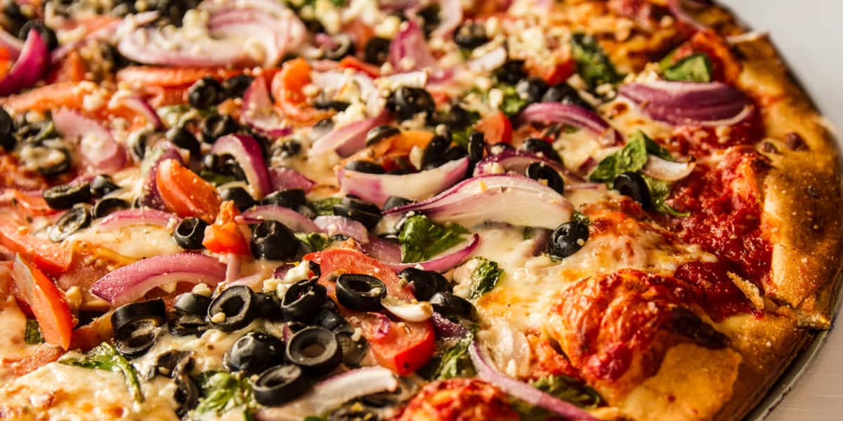Close-up of freshly baked vegetable pizza topped with black olives, red onions, tomatoes, spinach, and melted mozzarella on a golden crust.