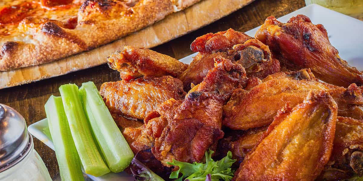 Plate of crispy buffalo-style chicken wings served with fresh celery sticks and dipping sauce, with a wood-fired pizza in the background at a casual restaurant bar.