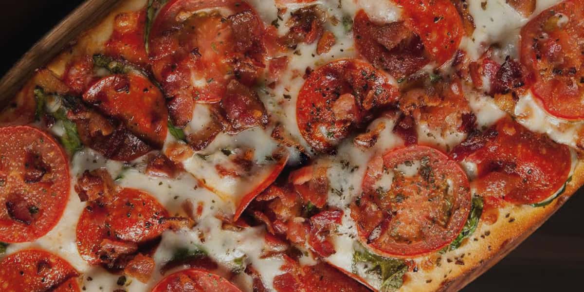 Close-up of loaded pizza topped with pepperoni, crispy bacon, sliced tomatoes, herbs, and melted mozzarella on a golden baked crust.