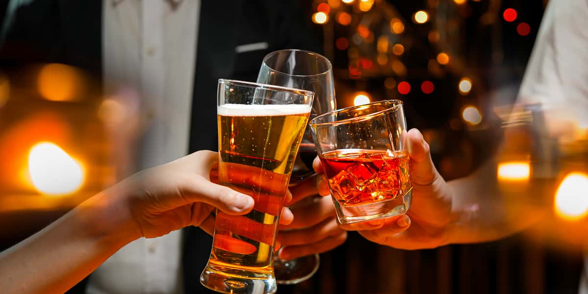 Guests clinking glasses of beer, red wine, and a whiskey cocktail in a warmly lit bar, capturing a celebratory nightlife moment with glowing candlelight ambiance.