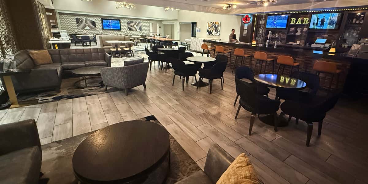 Spacious modern hotel bar and lounge with comfortable seating, round tables, large TV screens, and a full-service bar creating a relaxed social dining atmosphere.