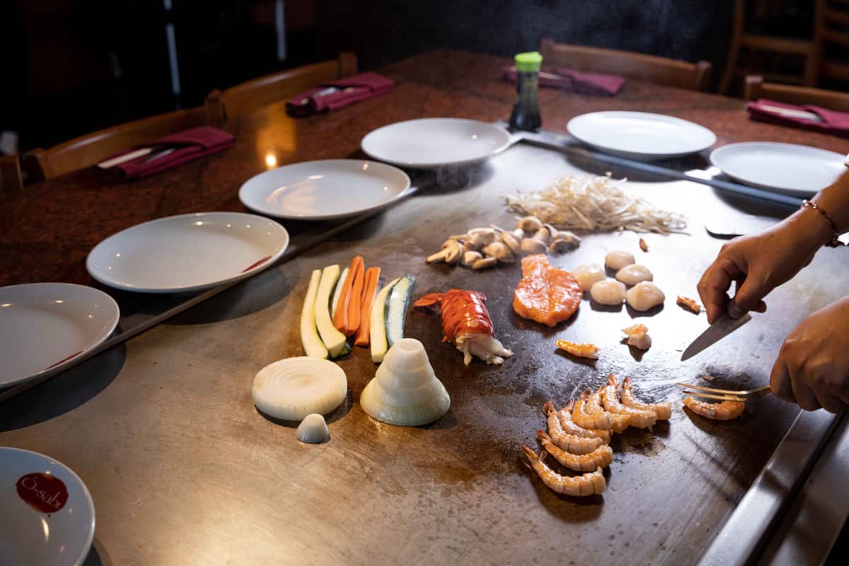A teppanyaki grill is set up for a live cooking experience, with a variety of fresh ingredients neatly arranged on the hot surface. Shrimp, sliced vegetables, rice, and sauces are spread out in preparation for cooking, while empty plates surround the grill, ready to be served. The scene captures the anticipation and interactive nature of teppanyaki dining, where guests watch their meal come to life right before them.