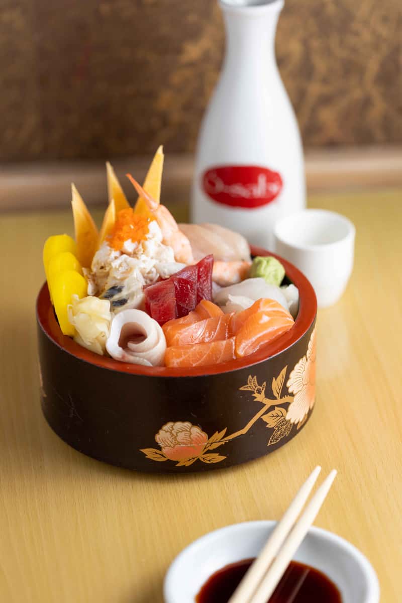 A beautifully presented Japanese-style seafood bowl sits in an elegant black lacquer bowl with gold detailing. The dish is filled with vibrant, fresh ingredients including slices of salmon, shrimp, squid, and other assorted seafood, all arranged over rice. Bright garnishes like lemon wedges and green leaves add color and freshness, while a delicate shrimp head is placed on top for a striking visual touch. In the background, a small white cup—likely for tea or sake—complements the refined dining setting.