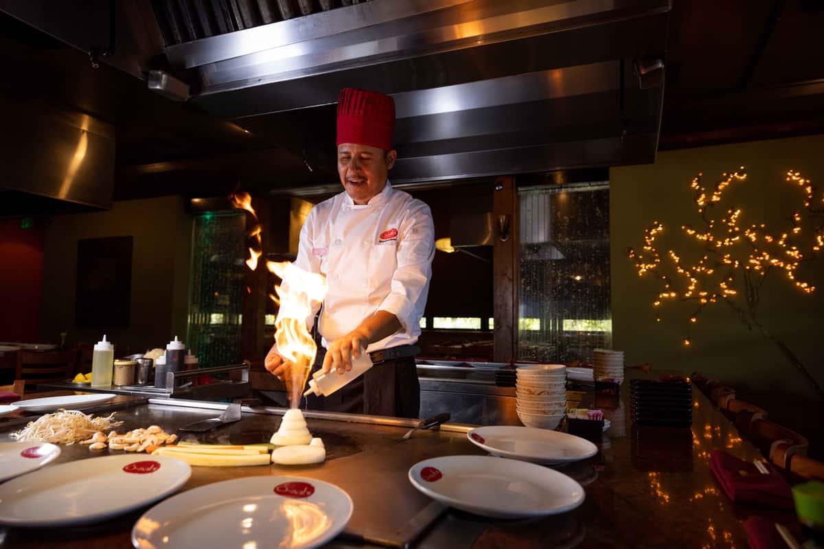 A skilled teppanyaki chef stands behind a flat-top grill, wearing a traditional red hat and white uniform. He is focused on preparing food with precision, using a spatula to cook ingredients right in front of guests. Flames rise from the grill, adding excitement and drama to the scene. In the foreground, neatly arranged plates and dipping sauces await the freshly prepared dishes, while the warm lighting and restaurant setting create an inviting and lively dining atmosphere.