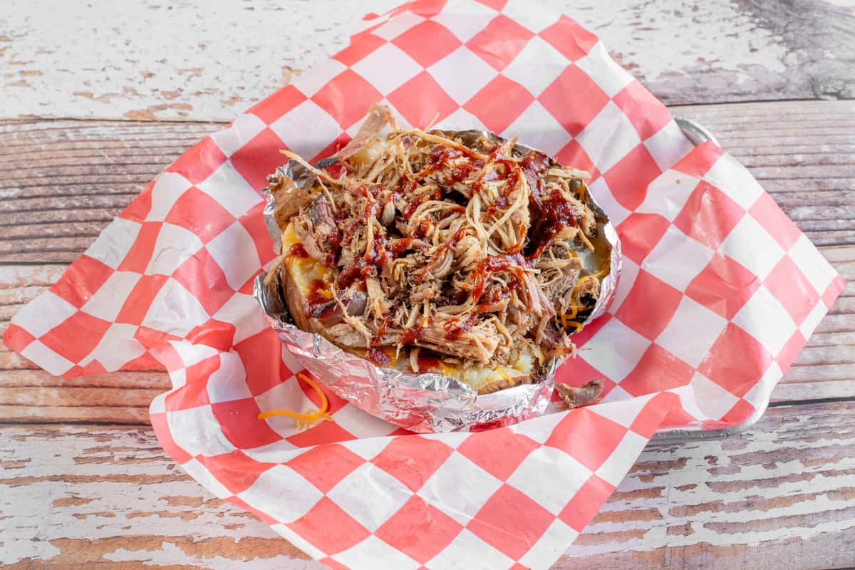 A loaded BBQ baked potato topped with pulled smoked meat, shredded cheese, and savory toppings, served on red and white checkered paper