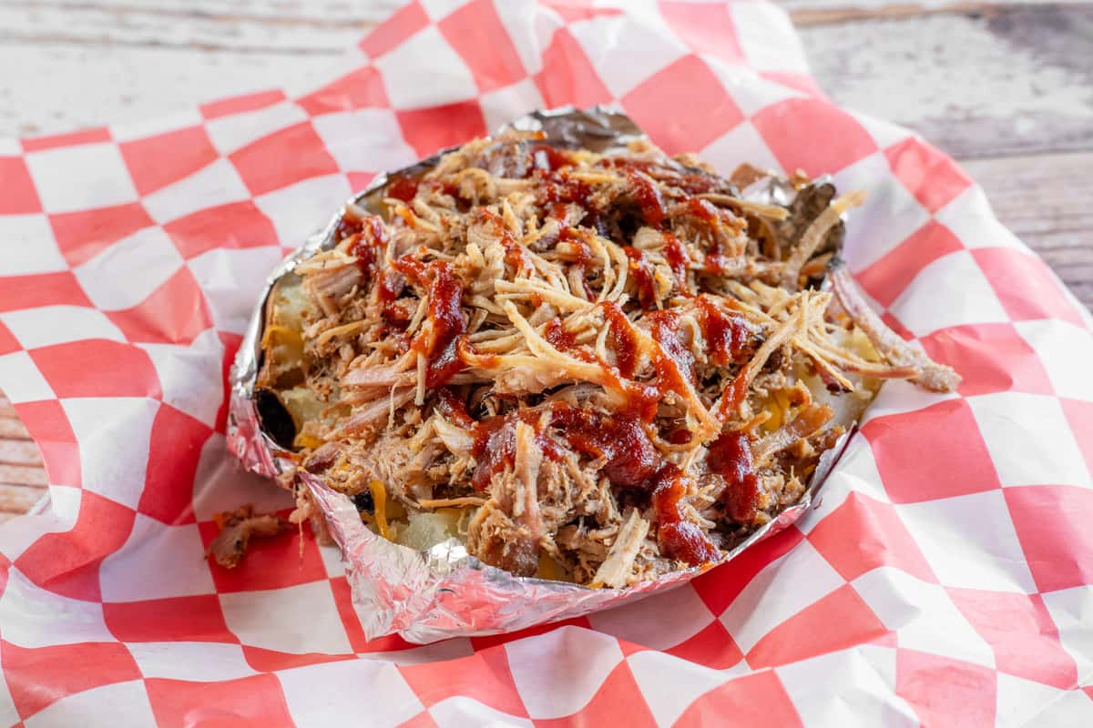 A loaded BBQ baked potato overflowing with pulled smoked meat, shredded cheese, and toppings, served in foil on red and white checkered paper