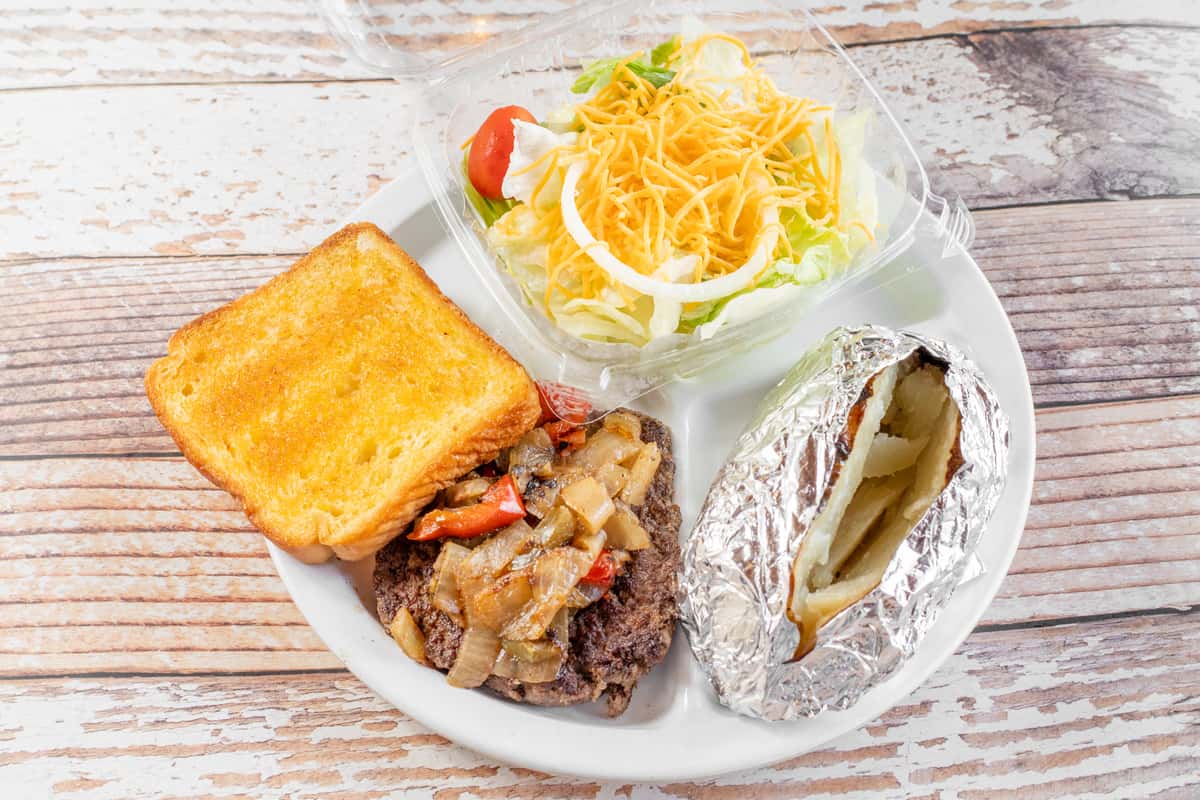 A hamburger steak plate with a smothered beef patty, cornbread, a baked potato, and sides of coleslaw and shredded cheese, served on a white plate