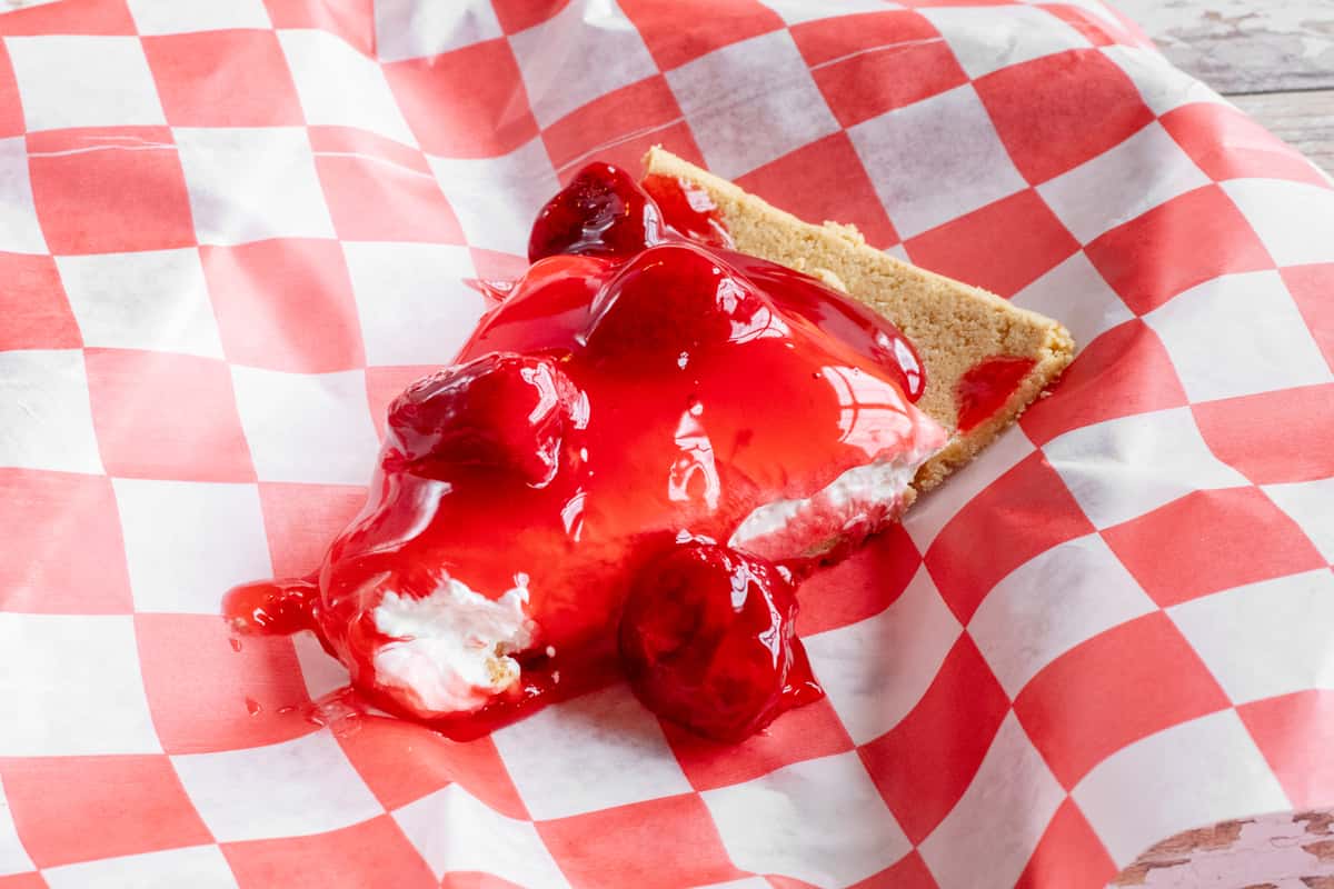 A slice of strawberry cream pie topped with glossy red strawberry sauce, served on red and white checkered paper