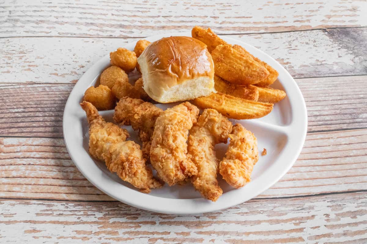 Crispy fried chicken tenders served with seasoned potato wedges, tater tots, and a soft dinner roll on a white plate.