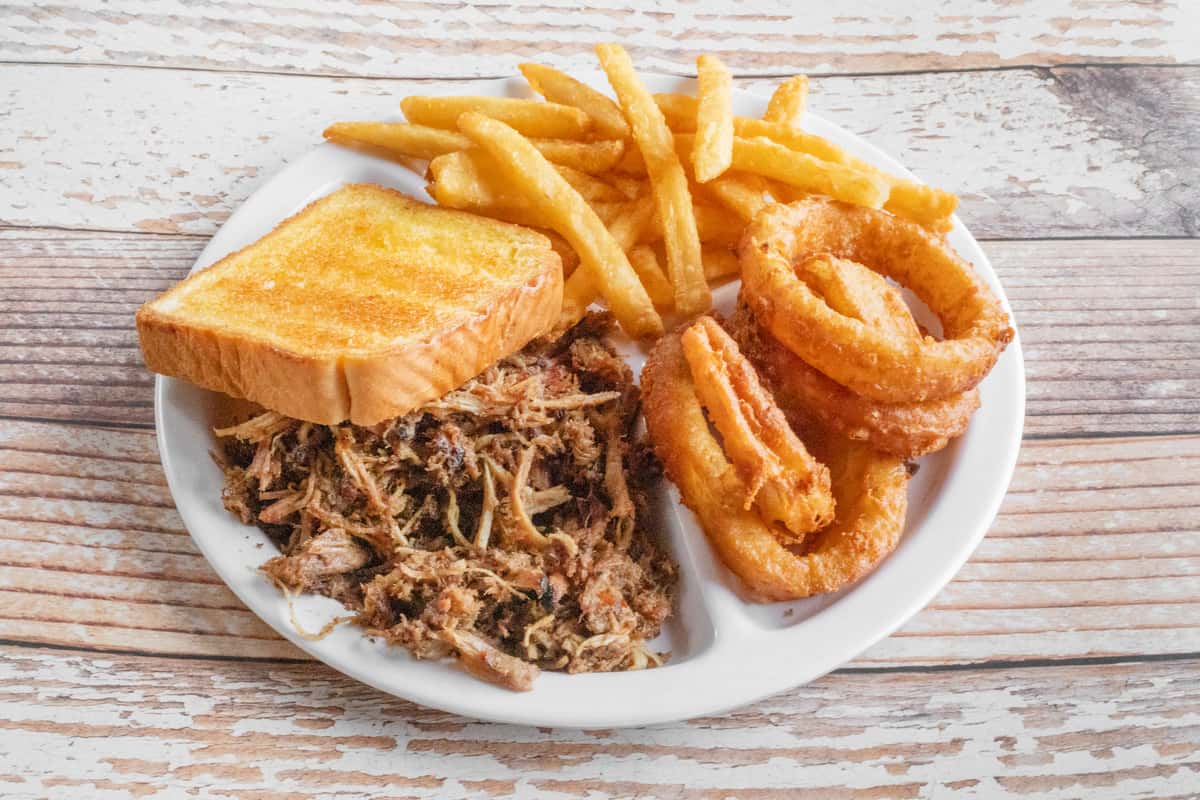 A BBQ plate with pulled smoked meat, golden french fries, onion rings, and cornbread on a white plate