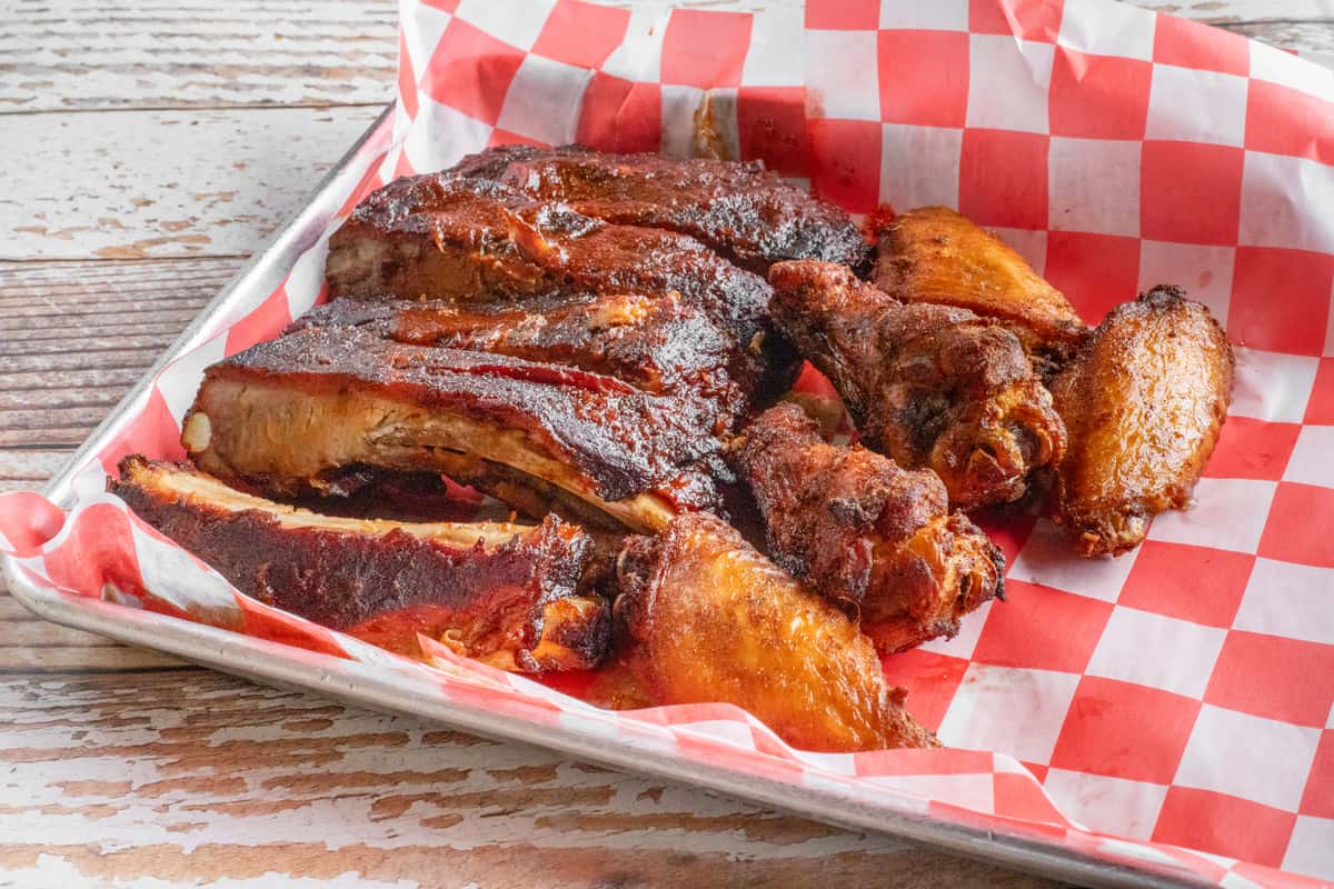 A large tray of slow-smoked BBQ ribs and glazed chicken wings, caramelized and charred to perfection