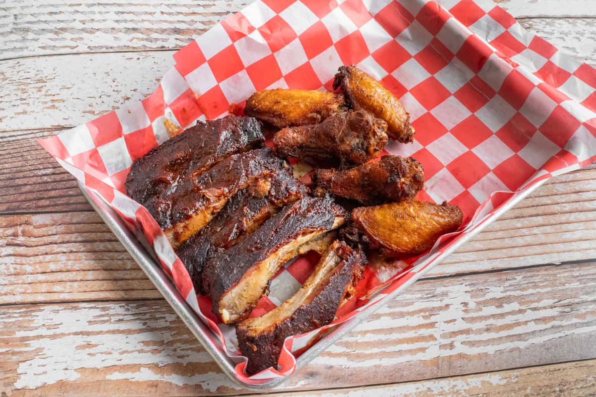 Slow-smoked barbecue pork ribs and crispy chicken wings served in a basket with red checkered paper and rich BBQ glaze.