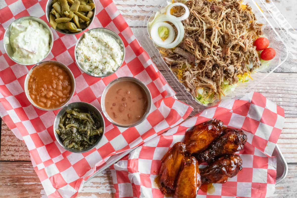 Southern barbecue spread with pulled pork salad, glazed chicken wings, baked beans, coleslaw, potato salad, green beans, and rice served on red checkered picnic paper.