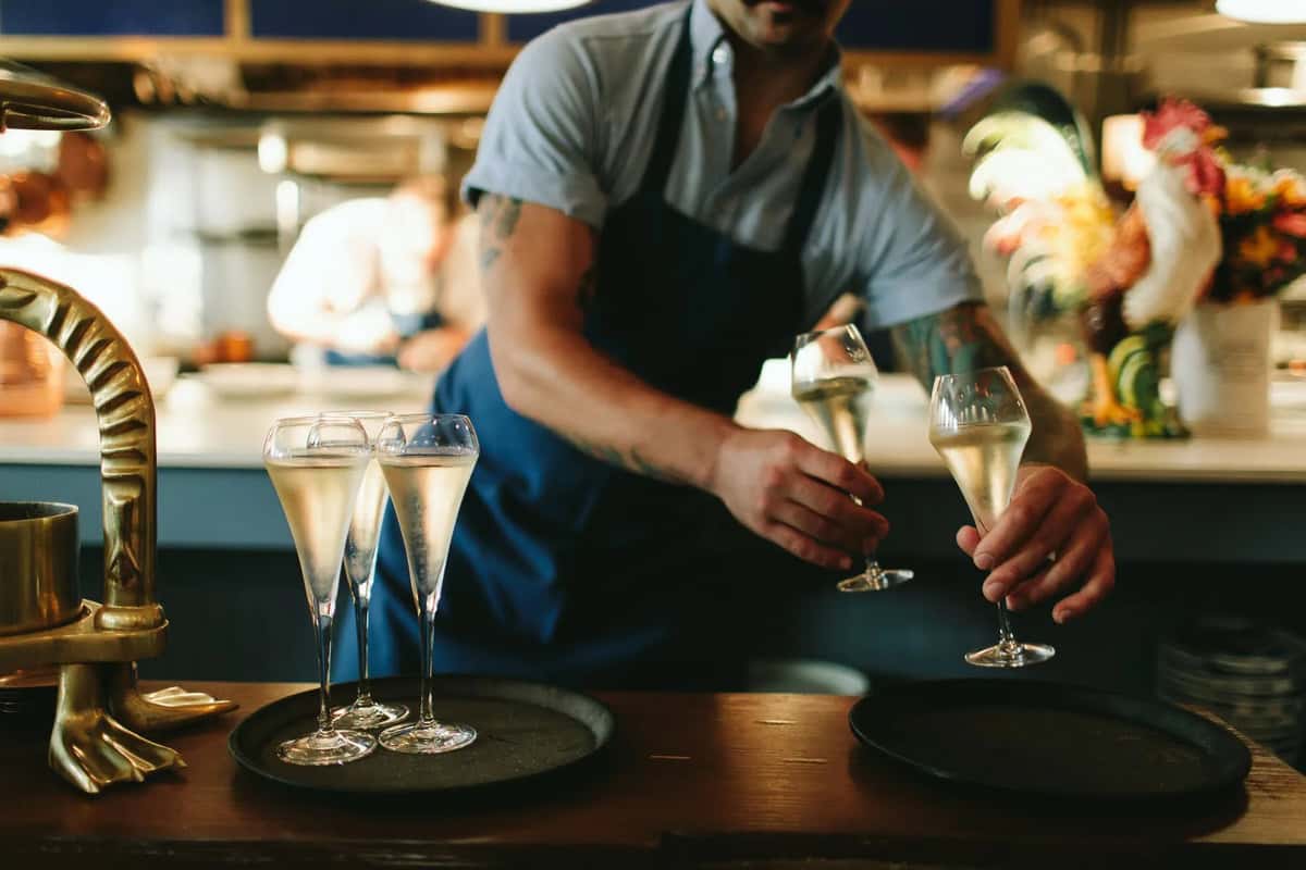 Server in a blue apron placing champagne flutes filled with sparkling wine onto a black serving tray in a restaurant.