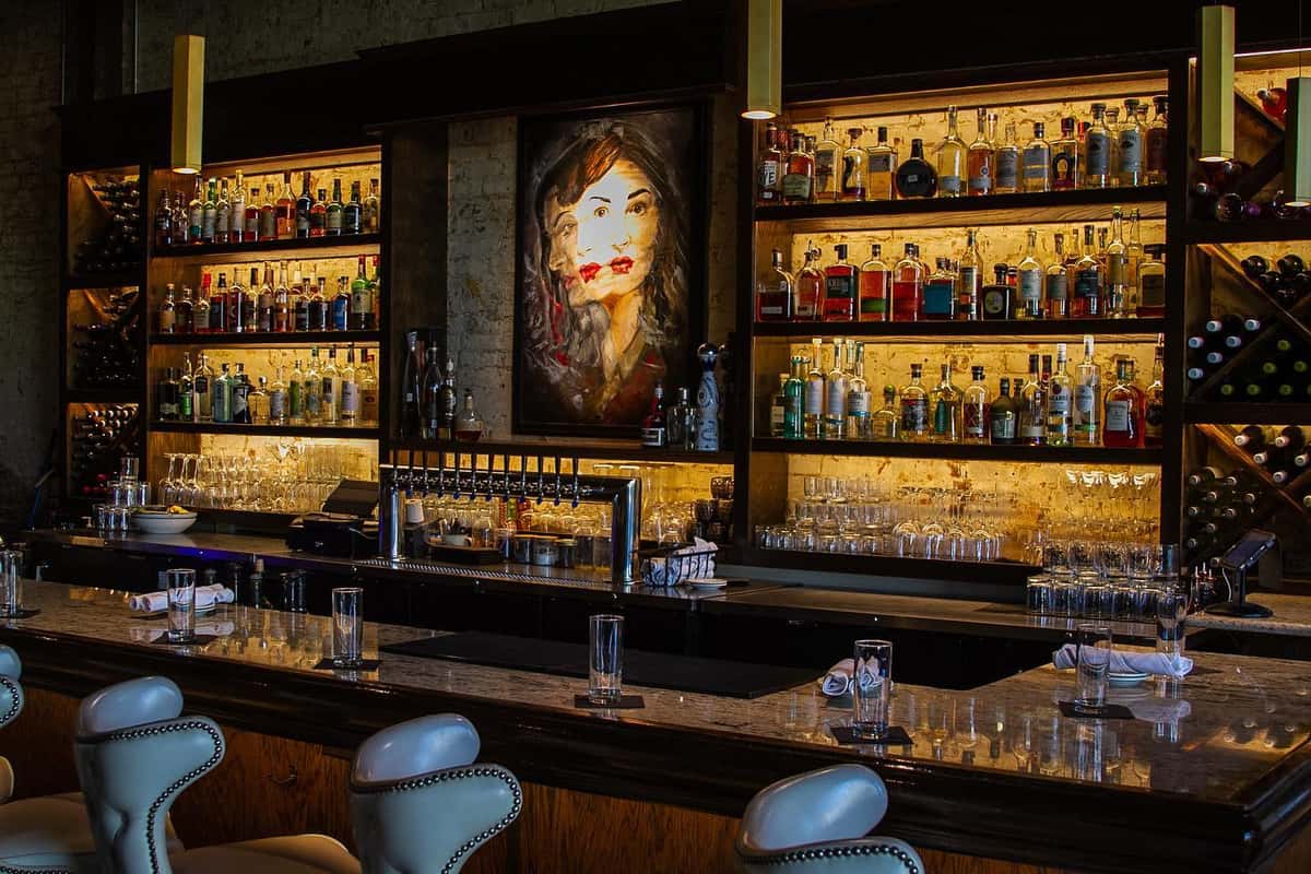 Elegant bar setup with marble countertops, leather barstools, backlit shelves of liquor bottles, and a bold portrait as the centerpiece.