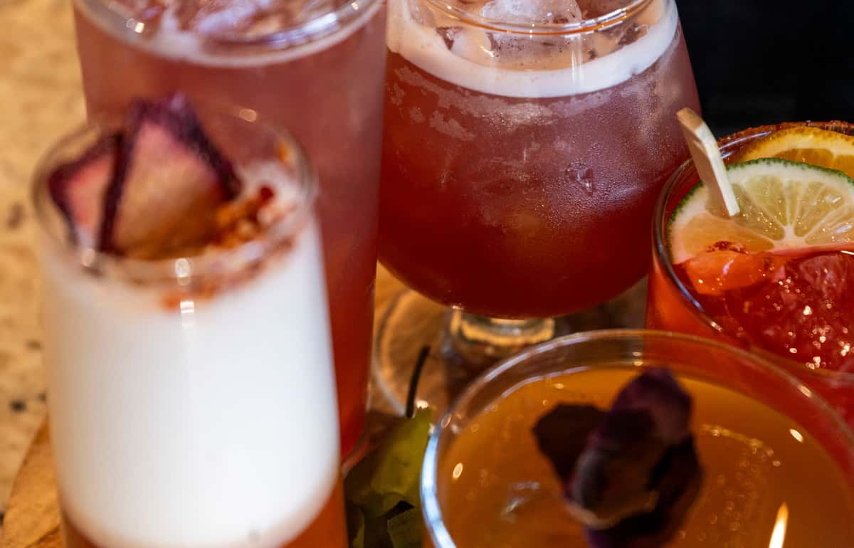 Assorted colorful cocktails garnished with citrus slices, berries, and foam served in a mix of glasses on a wooden board.
