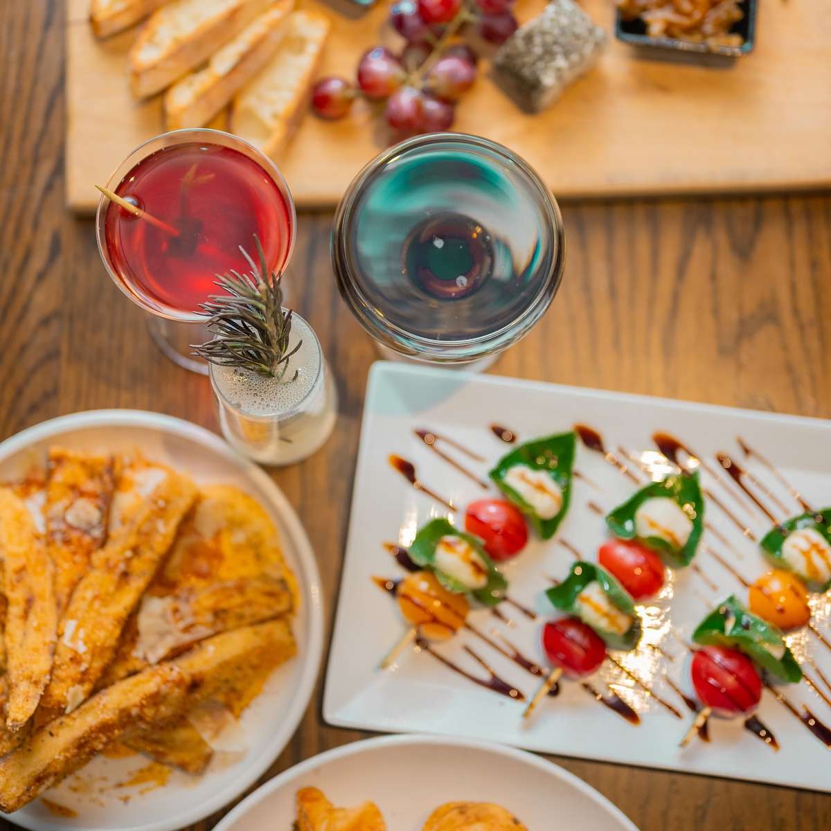 Colorful caprese skewers, cheese board with grapes and toast, and drinks served on a wooden table for a light appetizer spread.