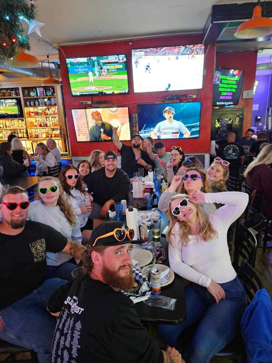Group of friends smiling and posing at a table inside The Upper Deck sports bar in Mount Airy, MD, enjoying drinks and game day atmosphere with big-screen TVs and a lively crowd in the background.