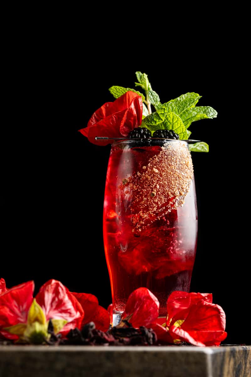 Bold red berry craft cocktail garnished with fresh mint, blackberries, and edible flowers against a dark background
