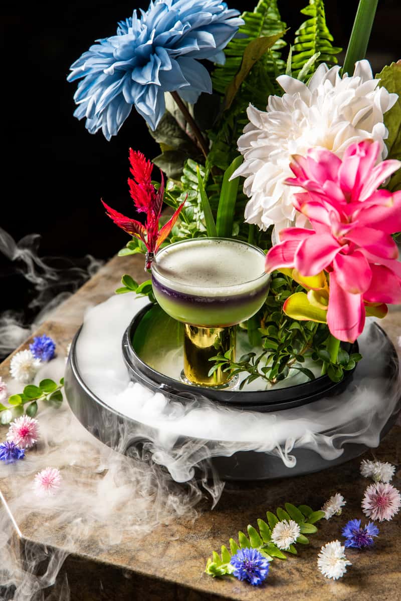 Artfully crafted floral cocktail with layered green and purple hues surrounded by tropical flowers and dramatic dry ice effects