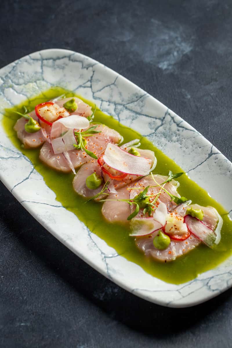 Fresh hamachi crudo served in vibrant herb oil with radish slices, chili, and microgreens on a modern ceramic plate