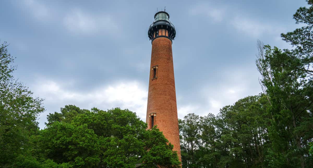 A tall brick lighthouse stands amongst lush green trees under a cloudy sky, not far from The Salty Rim, a fun beach restaurant.