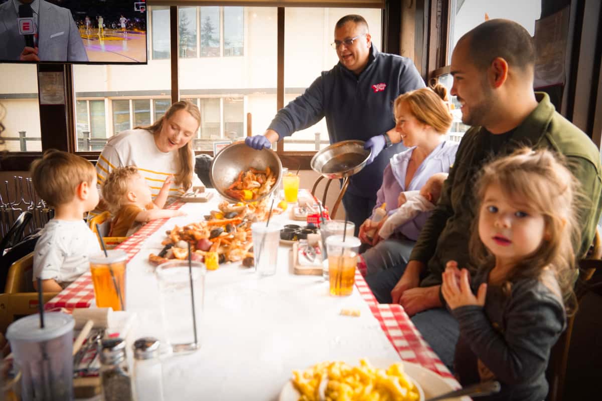 Image if two families sitting across from each other at a long table, with seafeasts spread infront of them. A crab pot staff member pours a seafeast on the table towards the center.