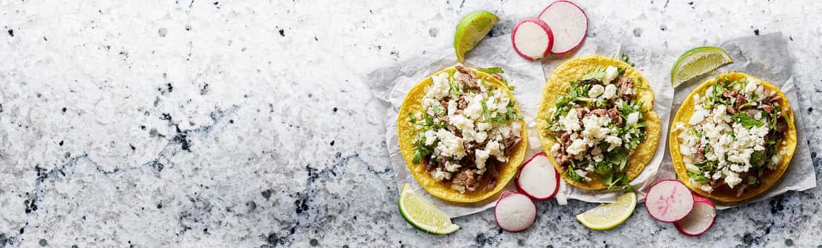 Three authentic street tacos on corn tortillas topped with chopped meat, crumbled cheese, fresh cilantro, and diced onions, garnished with sliced radishes and lime wedges on a marble surface.