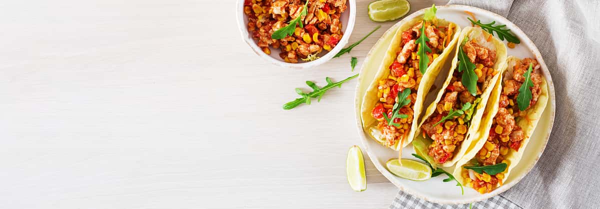 Three soft corn tortilla tacos filled with seasoned sautéed shrimp and corn mixture, topped with fresh greens, served with lime wedges on a light table setting.