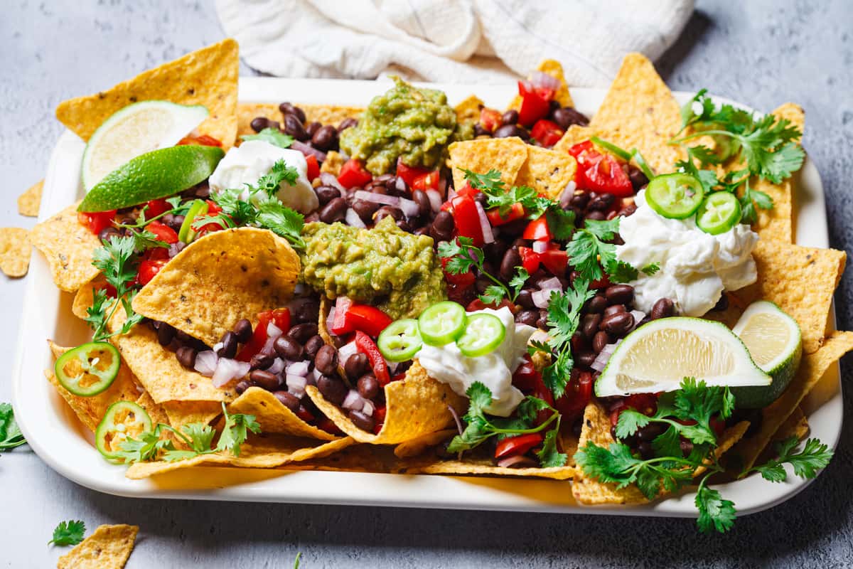 Loaded nachos topped with black beans, guacamole, sour cream, fresh pico de gallo, jalapeño slices, and lime wedges on a colorful Mexican restaurant platter.