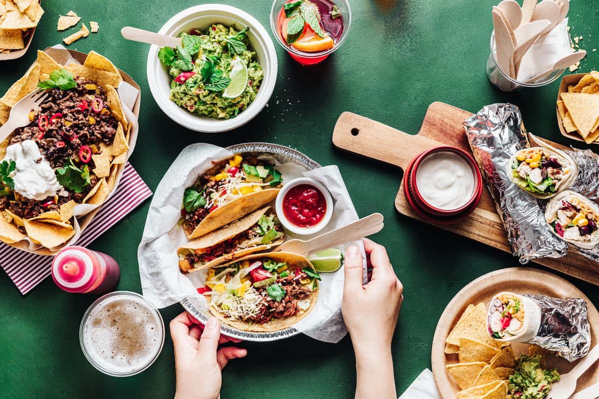 Colorful Mexican food spread featuring street tacos with seasoned meat, fresh cilantro, and cheese, served with tortilla chips, guacamole, salsa, and refreshing drinks on a vibrant green table.