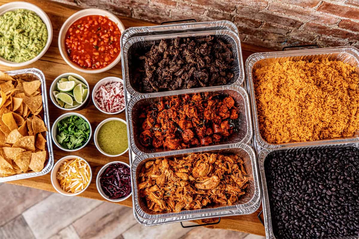 Mexican catering spread with trays of grilled meats, seasoned rice, black beans, and toppings like guacamole, salsa, cilantro, cheese, and tortilla chips on a rustic wooden table.