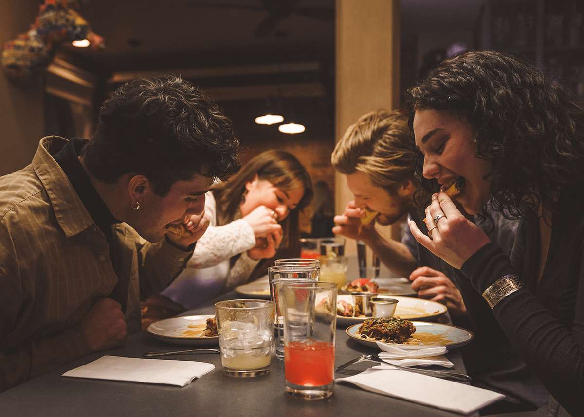 Friends enjoying tacos together at a cozy restaurant, leaning over plates of Mexican food with cocktails, capturing a warm, social dining atmosphere.