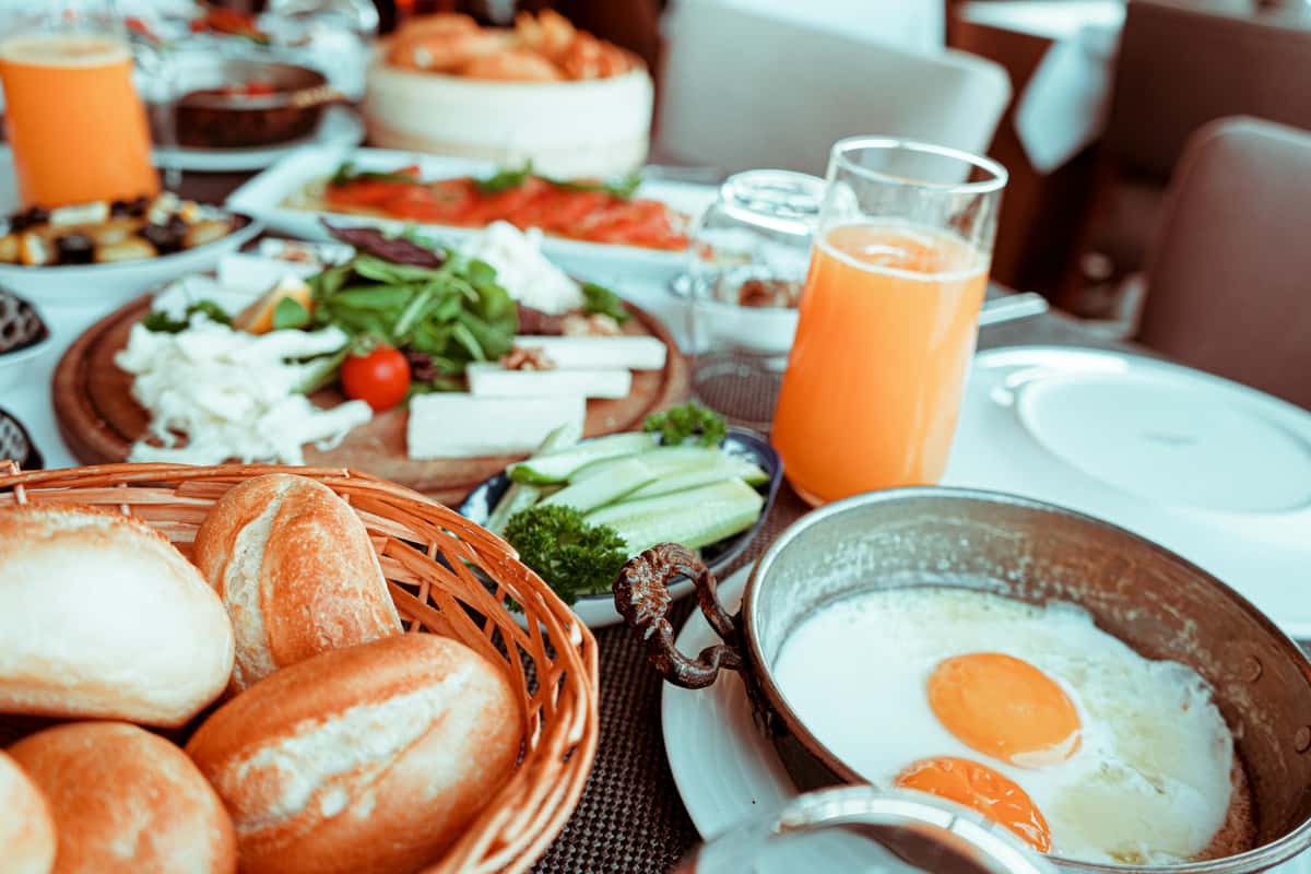 Breakfast spread with sunny-side-up eggs, fresh bread rolls, orange juice, cheese, cucumbers, and mixed greens on a dining table.