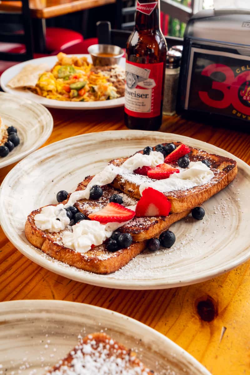 Golden French toast topped with whipped cream, red strawberries, and blue blueberries, served on a rustic wooden table with other breakfast dishes and a beer bottle in the blurred background.
