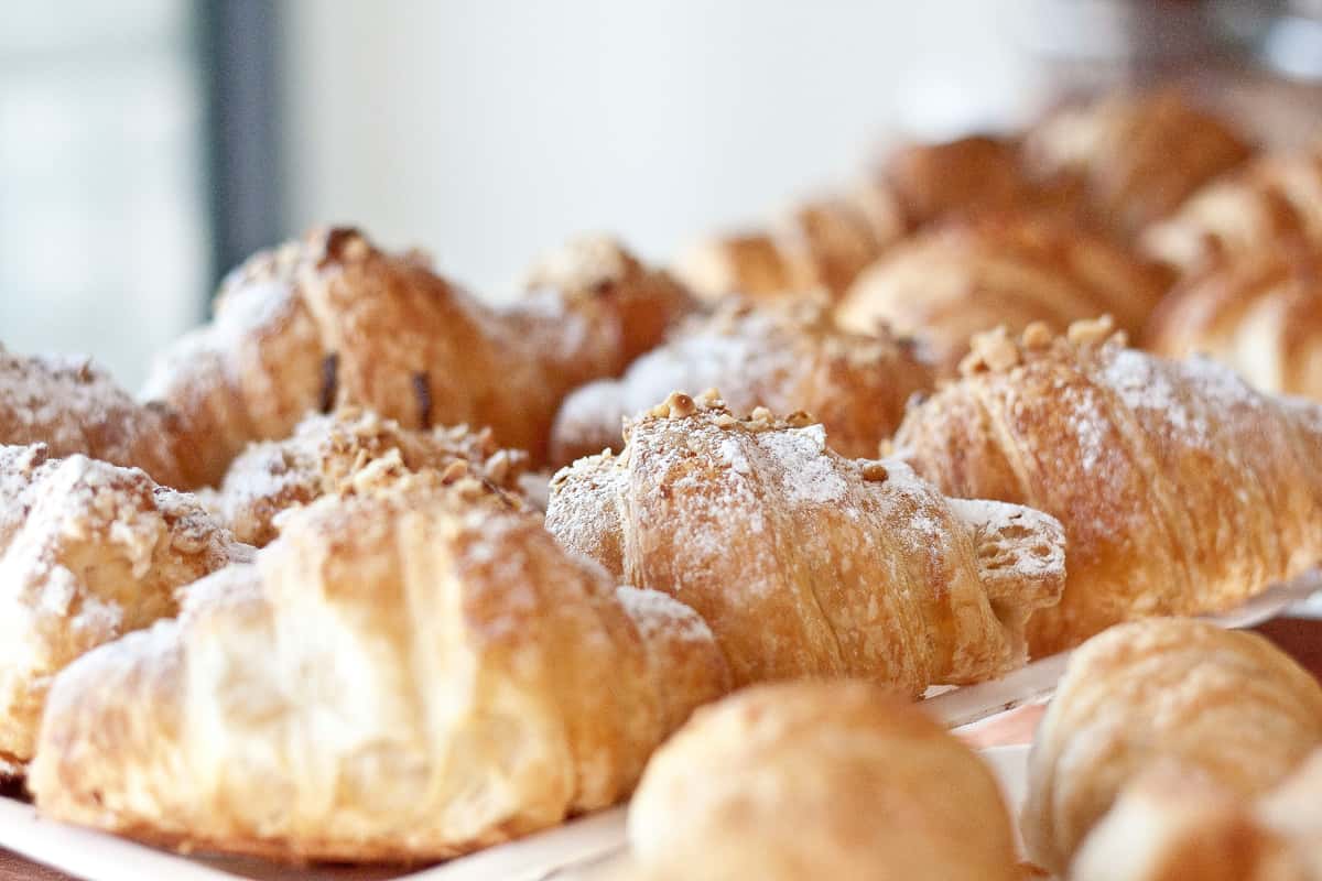A close-up of freshly baked croissants, some topped with powdered sugar and almond flakes, arranged neatly on a tray.