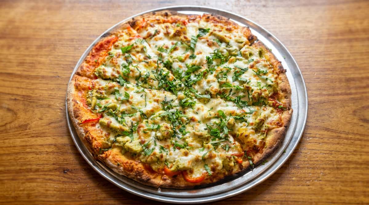 Pizza with melted mozzarella, pesto chicken, red sauce, and fresh basil, on a metal tray.