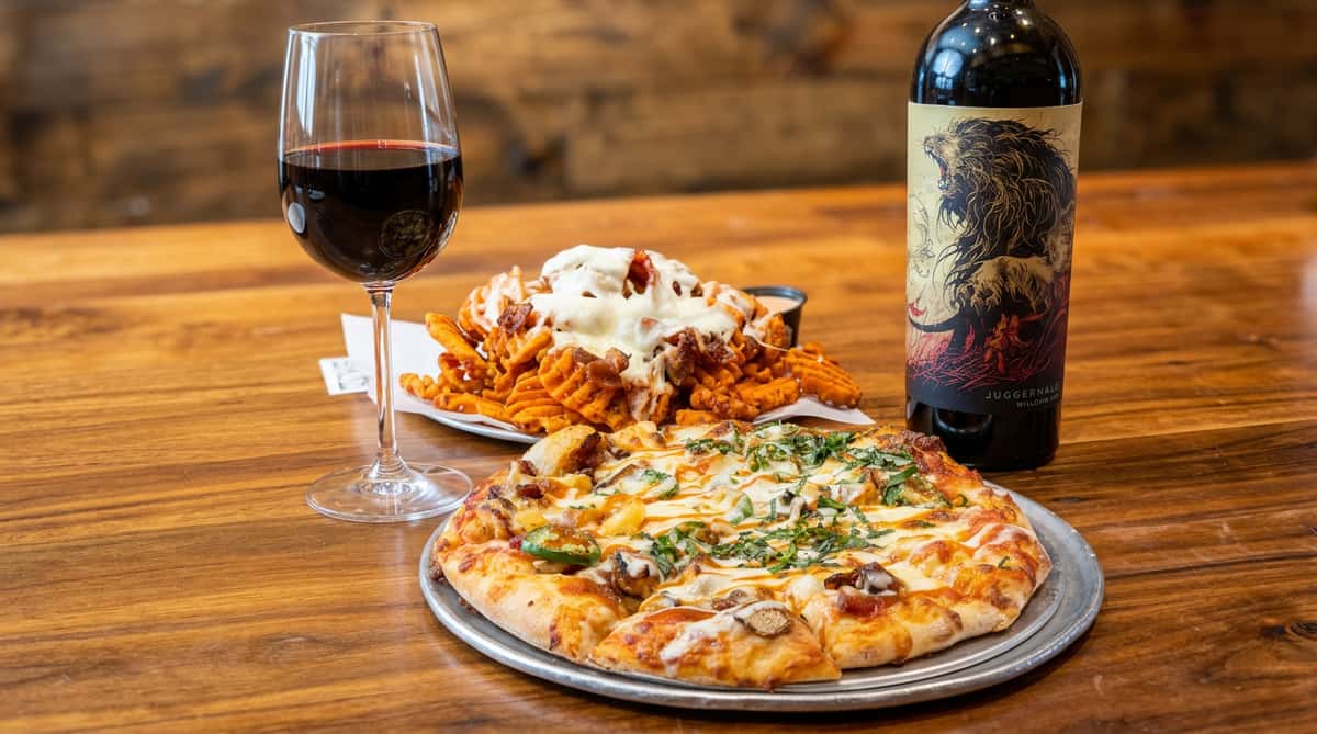 Pizza with jalapeños, mushrooms, and basil, a plate of waffle fries, a glass of red wine, and a bottle of red wine.