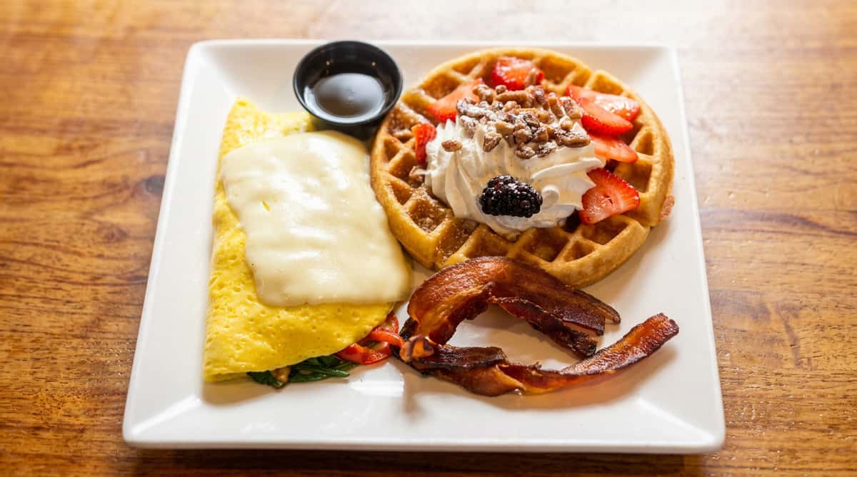 Breakfast plate with an omelet topped with cheese, a waffle with strawberries, whipped cream, and nuts, two slices of bacon, and a side of syrup.