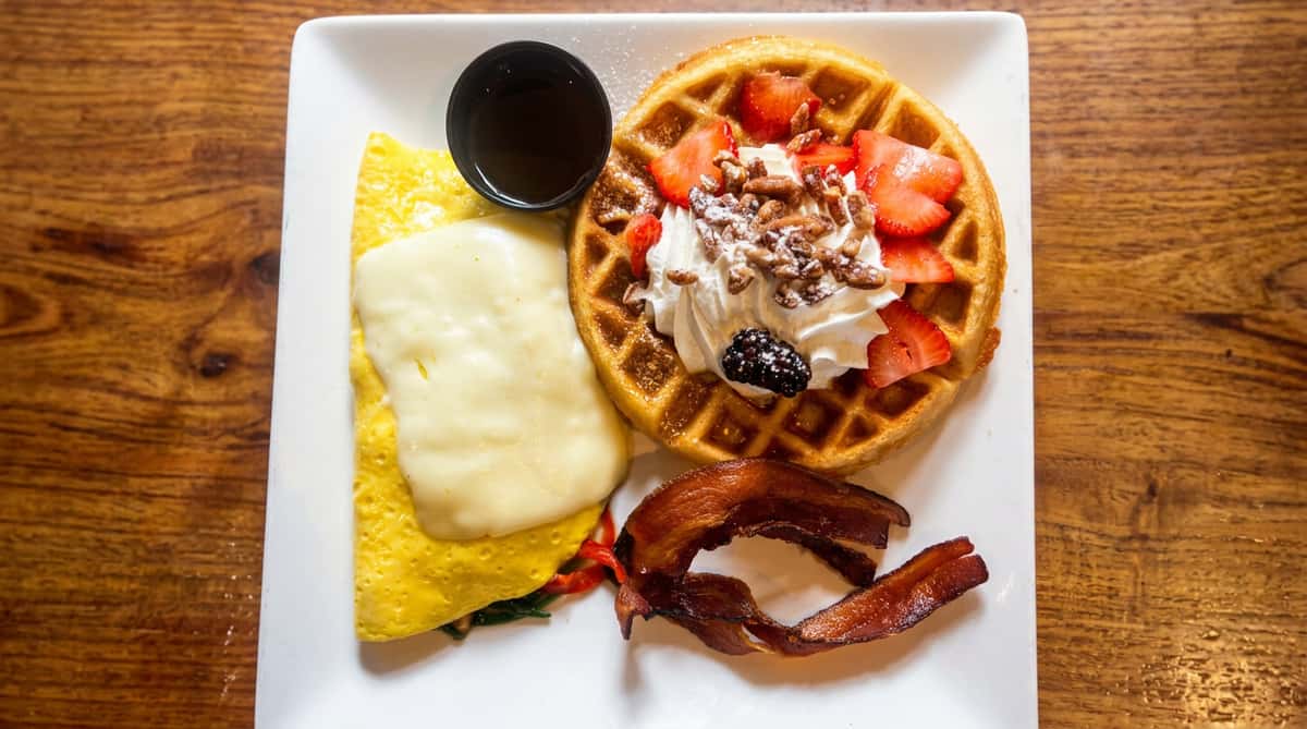 Breakfast plate with a waffle topped with strawberries, whipped cream, and nuts, alongside roasted potatoes and a cheese omelet.