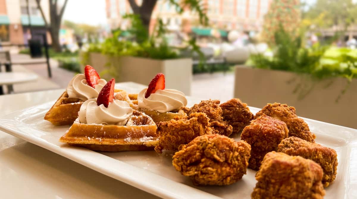 Chicken and waffles on a white platter, with whipped cream and strawberries, outdoors.