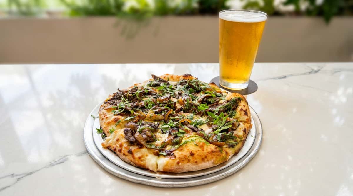 Pizza with dark meats, green pesto, white cheese, and parsley, next to a glass of beer on a marble table.