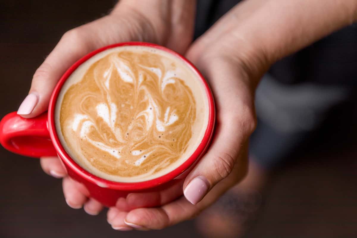 A warm latte with intricate foam art, served in a red cup and held in both hands.
