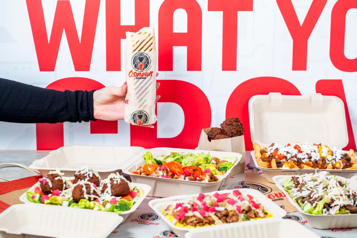 A variety of foods on a table and a hand holding a wrap from Osmow's Mediterranean
