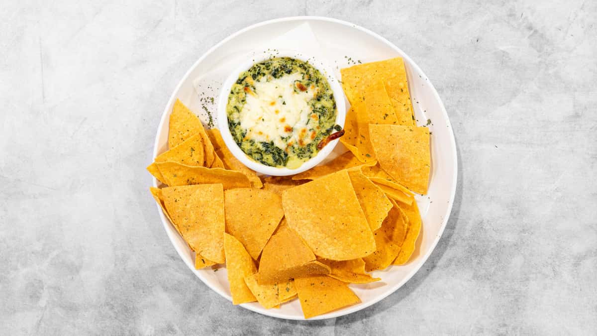 Coaches Corner's creamy green spinach dip with melted cheese, served with golden tortilla chips on a white plate.