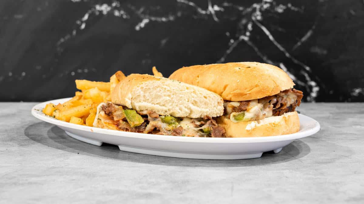 Coaches Corner's two cheesesteak sandwiches with melted cheese, green peppers, and golden fries on a white plate against a dark marbled background.