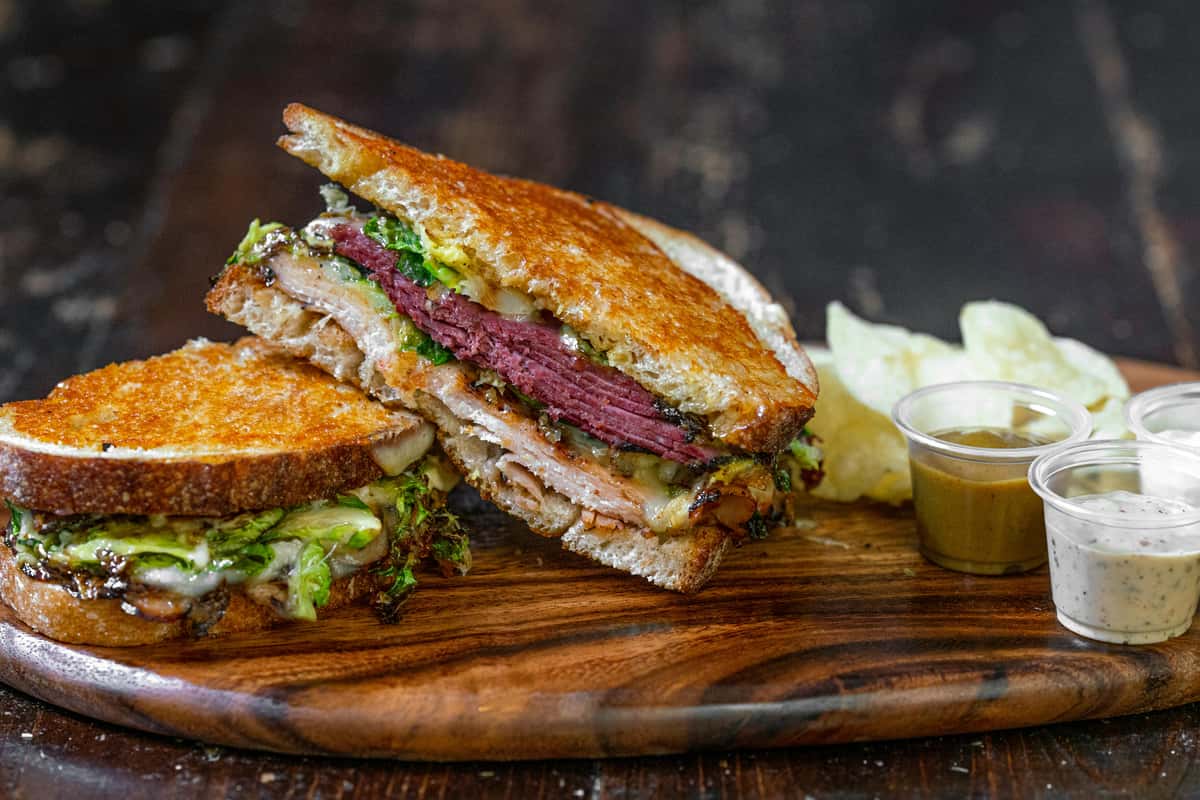 A grilled sandwich with layers of tender corned beef, melted cheese, and leafy greens on toasted rye bread, served with dipping sauces and chips.