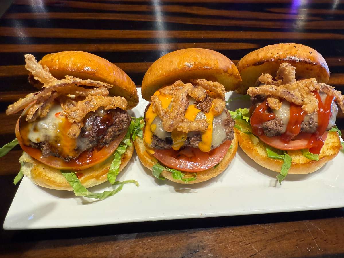 Three cheeseburger sliders topped with crispy fried onions, lettuce, tomato, and sauce on a white platter.